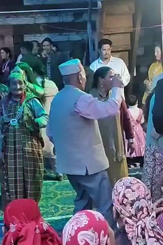 A group of people are gathered, some are dancing in traditional attire. The scene appears to be an outdoor or semi-outdoor event, possibly a celebration or cultural gathering, with wooden structures visible in the background and colorful decorations or lighting. The individuals are engaged in various activities, including dancing, watching, and interacting.