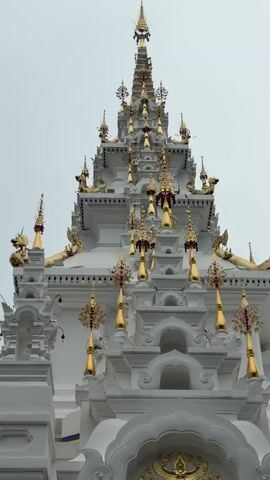 A close-up, vertical shot pans across the intricate white and gold facade of a temple. The camera begins by focusing on the topmost spire, adorned with golden finials and decorative elements. As the camera moves down, it reveals multiple tiered layers of the temple's roof and walls, all decorated with ornate golden carvings and pointed spires. Several hanging golden ornaments with red accents are visible. The lower portion of the facade features a large, golden emblem and an arched entryway. To the right of the entrance, a flagpole with a multi-colored flag (red, yellow, white, and blue) stands prominently. Further to the right, a small, dark sculpture of an animal-like creature is partially visible. The sky above is overcast and appears light gray.