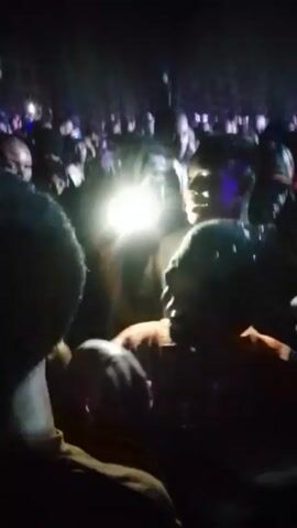 The video takes place at night amidst a crowd of people. Several individuals are visible, including a person with short, bleached hair wearing a white shirt and a chain. People are holding up phones to record. The crowd appears energetic, though the lack of visual clarity obscures specific actions and interactions. The environment suggests an outdoor event or gathering, with indistinct lights adding to the bustling atmosphere.