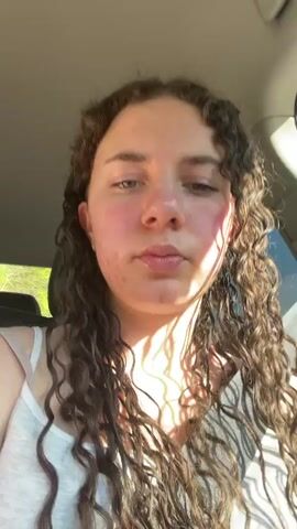 The video shows two people in a car. A woman with long, curly hair wearing a white tank top is in the passenger seat, facing the camera and holding up her hand in a rock-on gesture. She appears to be lip-syncing to music. The camera then shifts to a man driving the car. He is wearing sunglasses and a white shirt. The car is moving, and trees can be seen through the window.