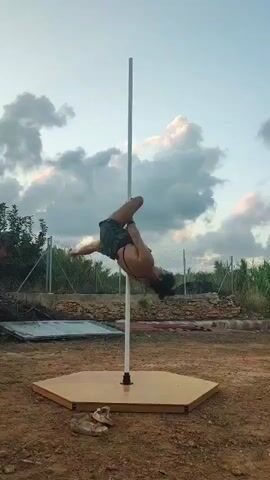 A person with dark hair, wearing a black halter top and dark shorts, is performing a pole dance routine outdoors. They are inverted, suspended from a white pole that is anchored to a hexagonal wooden base. Their legs are bent and wrapped around the pole, with their body in a curved position. The background shows a dry, outdoor environment with a fence, some vegetation, and a cloudy sky. There are a pair of sandals on the ground near the base of the pole. The person's head is tilted back.