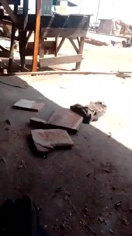 The video shows a view from under a dilapidated, open-air wooden structure, likely a market stall.  Outside the structure, there is a dirt or sand-covered open area. Other stalls or buildings are visible in the background, with some activity suggesting people are present. Debris and scattered objects are on the ground inside and just outside the structure. A large, dark-colored object resembling a bag sits in the foreground.  The video appears to be shot during the daytime with bright sunlight creating sharp shadows.