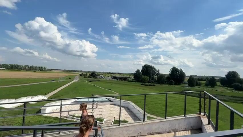 The video begins with a view of a white concrete structure with a metal railing. Two women are standing on this structure, facing away from the camera. The woman on the right has her hair in a bun, and the woman on the left has her hair down. They are both wearing dark-colored tops and light-colored bottoms. The video then pans to the right, revealing a wide, green landscape under a blue sky with scattered white clouds. The landscape features rolling hills, scattered trees, and a small body of water. In the distance, there are fields and more trees. The camera continues to pan, showing a path or road winding through the green fields and some small structures that appear to be buildings or pavilions. A small vehicle, possibly a lawnmower or golf cart, is visible on the grass in the middle distance. The video ends with a panoramic view of the landscape.