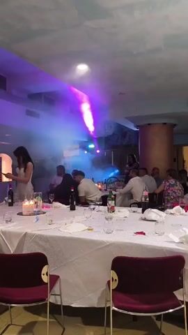 The video shows a gathering of people at what appears to be a formal event, possibly a wedding reception or a banquet. The scene is dimly lit, with colored lights and smoke effects creating a festive atmosphere. Several round tables are set up with white tablecloths, plates, glasses, and bottles of wine and water. In the foreground, two chairs with dark red upholstery are visible. A woman in a white dress is standing near one of the tables, looking at her phone. In the background, a group of people are dancing or socializing, with some individuals highlighted by blue and purple lights. Another group of people are seated at the tables. The overall impression is one of a lively celebration.