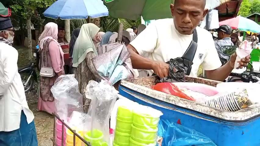 The video shows a street scene with a vendor selling ice cream from a cart attached to a motorcycle. The vendor, a man wearing a white t-shirt and a crossbody bag, is serving a customer, handing them an ice cream cone. Behind the vendor, there are several people, including women wearing hijabs and men wearing caps, suggesting a bustling market or gathering. Several umbrellas, in blue, green, and red, provide shade. In the foreground, plastic cups filled with colorful liquids, likely beverages, are visible on the cart. The environment appears to be outdoors, with trees and a building in the background. The overall atmosphere seems lively and busy.