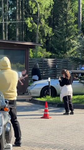 The scene shows a group of people near a silver car. A person in a yellow hoodie stands in the foreground, their back to the camera.  A woman in a black jacket and white top is standing next to the car, and a woman with dark hair is taking photos of the car with a phone. The silver car has black wheels and is partially elevated on wooden planks. There is an orange traffic cone in front of the car.  Behind the car is a wooden fence and beyond that, trees are visible.  A small building with a window and a black motorcycle are also visible in the scene.  The ground is paved with bricks. The scene is bathed in sunlight.