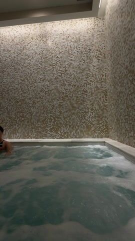 The scene is an indoor hot tub area. The hot tub itself is rectangular with bubbling, frothy water. The tub’s rim is trimmed with a white marble-like material. A person with short, dark hair and a swimsuit is partially visible in the corner of the hot tub. The hot tub is surrounded by tiled walls, which consist of small, mosaic-style tiles in various shades of brown and white. Above, there is a vent in the ceiling with a light fixture above it, illuminating the scene.