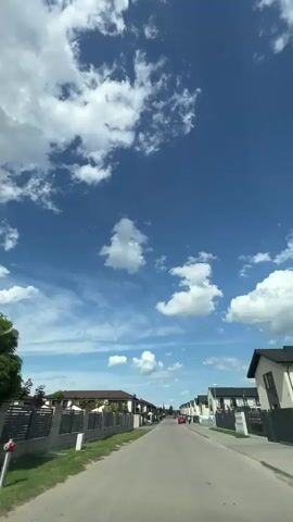 The video shows a view from a moving vehicle, looking down a paved residential street under a bright blue sky with scattered white clouds. The street is lined with modern-style houses on both sides. On the left, there are fences and houses with brown roofs and beige or white walls. On the right, there are similar houses, some with dark roofs. A few cars are parked along the street, including a silver car on the left and a red car further down on the right. A person in dark clothing is walking on the sidewalk on the right side of the street. The camera is positioned at a height consistent with being inside a car, with the dashboard or part of the car's interior possibly visible at the bottom edge of the frame. The overall impression is a sunny day in a suburban neighborhood.