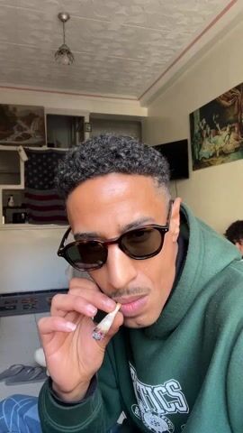 The video shows a man smoking what appears to be a marijuana cigarette. The man is light-skinned, wearing sunglasses and a green hoodie. He has short, curly hair. The man is in a room with artwork on the walls and an American flag. Another person is partially visible in the background. The man exhales smoke from the cigarette. Music can be heard.