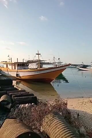 The video shows a harbor scene with several fishing boats docked. In the foreground, there are rusty barrels and fishing nets piled on the shore. A large yellow and white fishing boat is prominently featured in the middle of the frame, partially submerged in clear, shallow water. To the left, another green fishing boat is docked next to a concrete pier. The sky is clear with a few scattered clouds, and the sun is visible. The overall atmosphere is calm and serene, suggesting it is likely early morning or late afternoon. There are a few people visible on the pier in the distance, walking along the edge of the water. The scene appears to be a typical working harbor in a coastal area.