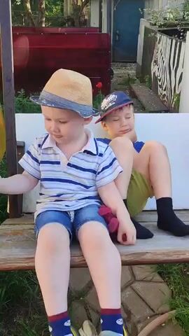 Two young boys sit on a wooden porch swing in a yard. The younger boy wears a tan straw hat, a striped shirt, denim shorts, and green Crocs. He holds a red stuffed animal. The older boy wears a baseball cap, a blue shirt, green shorts, and black socks. He yawns, resting his chin on his hand. The swing hangs in front of a white painted surface, with a red container and a blue door visible in the background. Green grass grows around the base of the swing, with scattered leaves and stones.