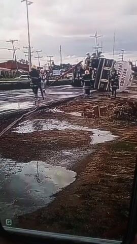 The video shows a capsized tanker truck with the word "PELIGRO" (Spanish for "danger") visible on its side. Firefighters in yellow helmets and dark suits are present at the scene. One firefighter is seen holding a hose, possibly spraying the area. The ground is wet and muddy, with puddles visible. Electrical poles and a building are in the background. The sky is overcast.