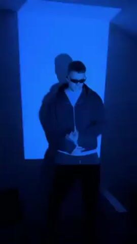 A man is dancing in a room with a red light, casting a shadow on the wall. He wears a black jacket, sunglasses, and a white shirt underneath. He gestures with his hands as he moves.