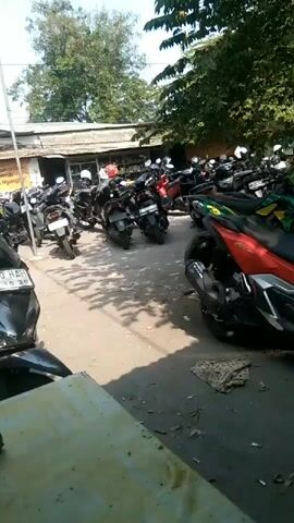 A parking lot is filled with parked motorcycles. Trees provide shade over the lot, and some buildings are visible in the background. The scene is captured from a low angle, focusing on the motorcycles. There are no people actively riding or interacting with the motorcycles.