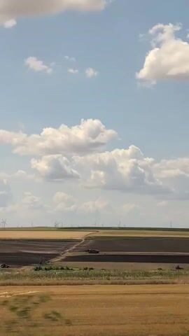 The video shows a landscape with a wind farm in the distance. The sky is blue with scattered white clouds. The foreground consists of fields of dry grass and plowed earth. A dirt road or path is visible winding through the fields. Several red vehicles appear to be working in the fields, possibly agricultural equipment. The wind turbines are spread across the horizon, their blades blurred by motion, indicating they are actively generating power. The overall impression is of a rural, open, and agricultural area.