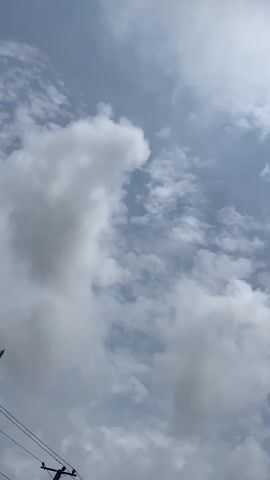 A vertical video shows a sky filled with clouds. The clouds are white and fluffy, with some appearing larger and more defined than others. The sky is a pale blue, and the lighting suggests it is daytime, possibly late morning or early afternoon. In the bottom left corner, a portion of a dark object, possibly a utility pole or construction equipment, is visible. There are no people or other discernible objects in the frame.