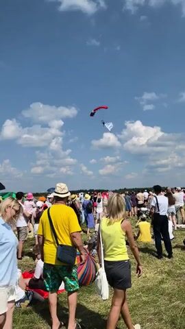 A wide shot captures a sunny outdoor event with a clear blue sky dotted with fluffy white clouds. In the foreground, a crowd of people stands on a grassy field, facing away from the camera. They are casually dressed for warm weather, with some wearing hats. In the middle distance, a skydiver descends under a red parachute, holding a white banner. The skydiver is in the process of landing or maneuvering. Behind the skydiver, more people are visible, and beyond them, some tents and trees can be seen, suggesting the event is taking place in a park or open field. The overall atmosphere appears to be festive, with attendees looking up at the skydiver.