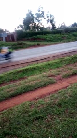 The video shows a road running vertically through a landscape, flanked by green grassy areas on either side. To the left of the road, there are trees and a structure partially visible, appearing to be made of concrete blocks. Adjacent to the road on the right is a reddish dirt path running parallel. The sky is overcast and bright. The perspective suggests the shot was taken from an elevated position.