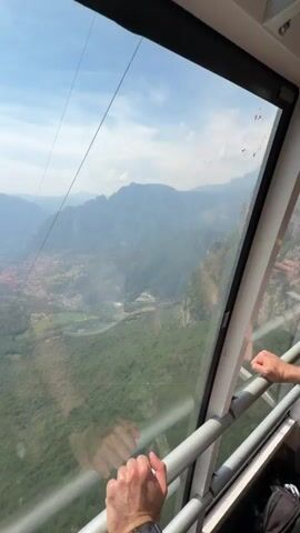 The video shows a view from inside a cable car ascending a mountain. The person in the cable car is holding onto the metal railing. Through the window, we see the landscape below, including a town nestled in the valley, green forests, and a lake in the distance. As the cable car ascends, we see the rocky, forested mountain side, and another cable car going in the opposite direction. The sky is blue with some clouds.