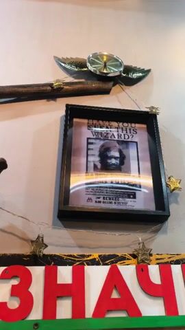 The media file is a short video, likely a snippet from a longer recording, showing a wall decorated with various items. The central focus is a framed "Undesirable No. 1" poster from the Harry Potter universe, featuring a picture of Harry Potter. To the left of the poster is another framed wanted poster, this one depicting the Joker from the Batman franchise. Above these posters, a decorative object resembling golden wings with a clock in the center is mounted on the wall. Below the framed posters, a colorful sign with large red Cyrillic letters that spell out "ЗНАЧ" is visible. The overall setting appears to be an indoor space, possibly a room or a shop, with a neutral-colored wall as the backdrop. The lighting is artificial, suggesting it could be daytime or nighttime. The decorations suggest a theme, possibly related to pop culture or fantasy, and the presence of Christmas-themed star lights strung across the wall adds a festive touch. No specific activity or event is taking place beyond the static display of these objects. The location context provided is Sterlitamak, Russia, which, while not directly evident from the visual content, provides a geographical frame of reference.