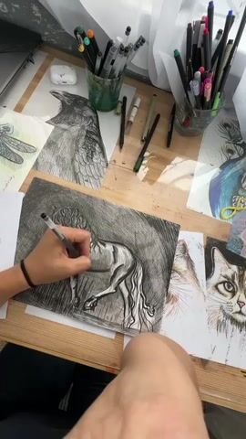 A high-angle medium shot shows a person's arm and hand drawing a horse with a pen on a piece of paper. The paper is on a wooden table, which is cluttered with art supplies, including a laptop, two cups filled with pens and markers, and several other drawings of animals. The drawings include a dragonfly, a crow, a peacock, and a cat. The lighting suggests it is daytime, with sunlight coming from a window on the right side of the frame. The person's lower leg and knee are also visible in the bottom right corner of the frame.