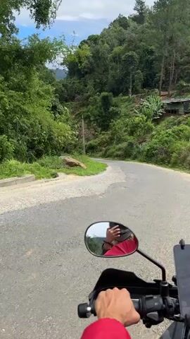 The video begins with a first-person perspective of someone riding a motorcycle on a paved road that winds through a lush, green, mountainous landscape. The road has some rough patches. The rider's hand, wearing a red sleeve, is visible on the handlebars. In the motorcycle's rearview mirror, a reflection of the rider is seen, holding a phone to take a selfie. The surrounding environment is characterized by dense vegetation, including trees and bushes, with a blue sky and white clouds visible above. The road appears to be in a rural or remote area, with a telephone pole visible on the left side of the road. The motorcycle is moving forward along the road, which curves to the right.