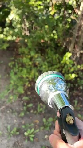 A hand holds a hose nozzle that is spraying water in a fine mist. The nozzle is silver and has green accents. The background is blurred and appears to be a garden or wooded area with green foliage and brown dirt. The water spray is catching the light, creating a shimmering effect.