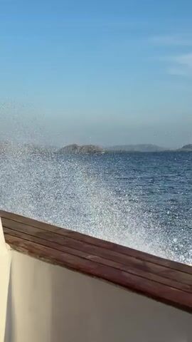 The video depicts a view from a boat moving on the water, with waves crashing against the side. A wooden railing is visible in the foreground. The water is choppy and a large spray of water is visible, obscuring the view. In the distance, there are land formations under a bright blue sky.