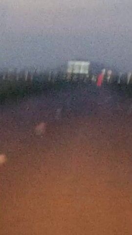 The video shows a blurry, low-resolution view from a moving vehicle. The scene appears to be a rural or semi-rural landscape, possibly at dusk or dawn, with a gray sky. In the distance, a prominent hill or mountain is visible. The hillside is dotted with lights, some appearing white and others red or orange, suggesting artificial lighting from buildings or vehicles. The foreground shows a dark road with faint white lines, indicating lane markings. The overall impression is of a brief, out-of-focus glimpse of a landscape with scattered illumination.