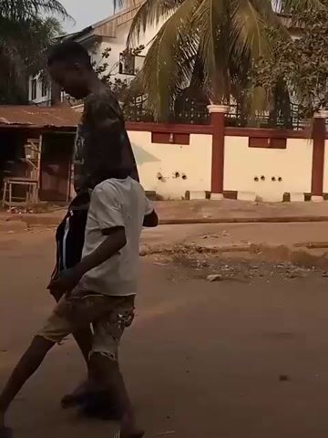 Two young males are on a dirt road. The older one wears a camouflage-patterned shirt and black shorts, and the younger boy has a white shirt and patterned shorts. The older boy balances a soccer ball on his foot. They stand next to each other, then walk down the dirt road together. Other buildings and people are in the background.