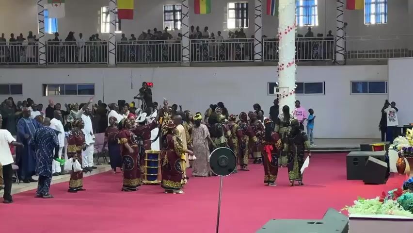 A group of people are dancing and singing in a large hall. They are dressed in colorful traditional clothing. The hall is decorated with flags and banners. There is a crowd of people watching the performance.