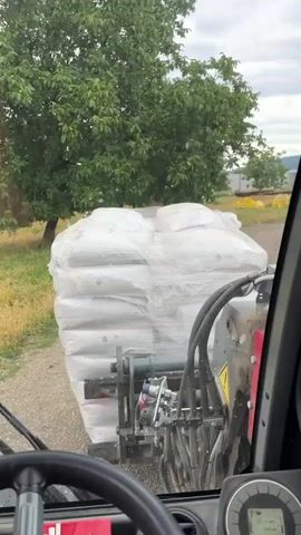 The video shows the interior of a vehicle, possibly a forklift or telehandler, with a pallet of large white bags covered in plastic wrap being carried on the front. The pallet is positioned in front of the vehicle's cab, obscuring some of the view. Through the windshield, a green tree is visible to the left, and in the background, there is a dirt road and some farm buildings. A digital display on the dashboard shows various indicators, including a level gauge and a load meter. The steering wheel and some tools are also visible in the foreground. The overall impression is of agricultural or industrial work being carried out in an outdoor setting.