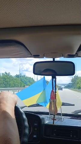 The video is filmed from inside a car, looking forward through the windshield. A person's hands are visible on the steering wheel, indicating they are driving. Two Ukrainian flags hang from the rearview mirror, obscuring a portion of the driver's face reflected in the mirror. In front of the car, on a highway, a large white truck with an orange concrete mixer drum is passing. The truck has text on its side, including "BETOH" and a phone number. Further ahead on the highway, other vehicles are visible. The environment outside appears to be rural, with trees and a cloudy sky. The video appears to be a short clip of a car journey.