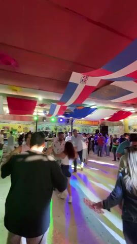 The video shows people dancing in a crowded indoor space. Numerous Dominican Republic flags are draped across the ceiling. A sign that says "COCO BONGO BAR" can be seen in the background. There are lights on the dance floor, and people are dressed in casual clothing. Some people are dancing in pairs, while others are standing and watching. The video is shot from the perspective of someone standing in the crowd.