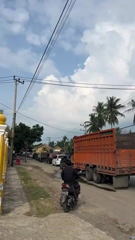 The video shows a street scene with several vehicles, including a large orange truck, a white minivan, a white car, a black SUV, and a motorcycle. The truck is the most prominent object, taking up a significant portion of the frame. It appears to be hauling wood. The vehicles are on a dirt road that seems to be undergoing some construction or repair, as evidenced by the rough surface and puddles. There are power lines overhead, and palm trees are visible in the background, suggesting a tropical or subtropical location. A building with the signage "SHOWROOM SUKSES LARIS MOTOR" is also visible, indicating a car dealership. A person is riding the motorcycle and appears to be interacting with someone on the side of the road. The sky is partly cloudy. The overall impression is that of a busy street in a developing area.