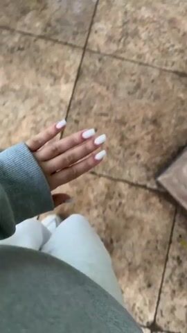 The video shows a first-person perspective of someone walking. The person is wearing a grey hooded sweatshirt and light-colored pants. They have white painted fingernails. The floor appears to be tiled with a brown and beige pattern. The video is shaky and slightly blurred, indicating movement. In the foreground, a hand is visible with painted nails. The hand moves slightly, and the camera shakes. In the background, the tiled floor continues. There is a glimpse of a dark rectangular object on the right side of the frame, out of focus.