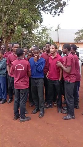 Here is a description of the video:

The video shows a group of teenage boys standing in an outdoor area with reddish-brown dirt. Most of the boys are wearing maroon shirts with a school logo, black pants, and black shoes. Some boys are wearing blue sweaters or shirts instead. They appear to be talking and socializing, and some are smiling or looking around. In the background, there are trees, a building with a gray roof, and parked cars. The atmosphere seems casual and informal.
