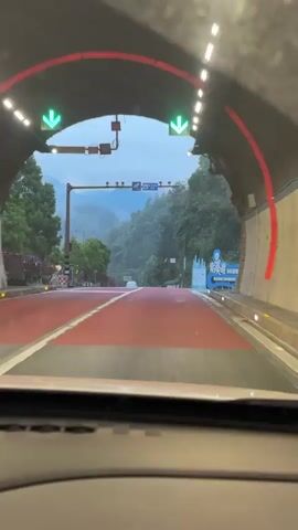 The video shows a view from inside a car as it exits a tunnel onto a red-paved road.  The road leads towards a mountain range shrouded in mist. Trees line the roadside. A white car passes on the left, and another car is visible in the distance ahead. A sign in Chinese with English translation, possibly welcoming visitors to a tourist resort, stands to the right of the road. The sky is overcast.  The windshield appears slightly wet.