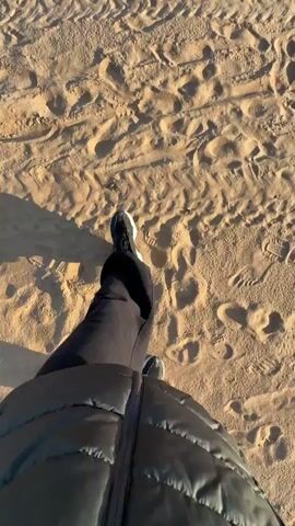 A first-person perspective video shows a person walking on a sandy beach. The person is wearing black pants and black sneakers. The sand has tire tracks and footprints. The video pans up to show the ocean, which is covered in green seaweed, and a blue sky with white clouds.