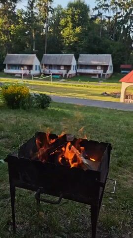 A close-up shot shows a metal barbecue grill filled with burning logs. In the background, several small, identical white cabins with wooden porches are visible, arranged in a row. A well-maintained lawn stretches between the cabins and the foreground grill. Trees form a dense backdrop to the scene. The lighting suggests it is either early morning or late afternoon.
