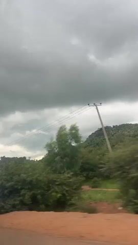 The video shows a view from a moving vehicle. The sky is overcast with dark, heavy clouds. In the middle ground, there is a lush green, tree-covered hillside or mountain. Power lines stretch horizontally across the frame, supported by utility poles. The foreground consists of a reddish-brown dirt road. The motion blur indicates the vehicle is traveling at a moderate speed.