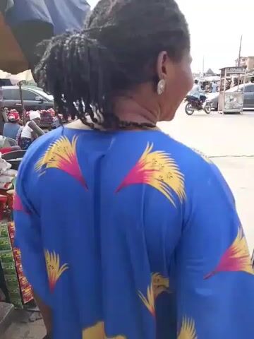 A woman in a blue, patterned dress is standing on a street corner in what appears to be an urban area. She turns to speak with another woman wearing a black, yellow, and white striped shirt. The street is paved with concrete blocks and has several motorbikes and cars driving by. There are buildings and shops visible in the background. The sky is overcast.