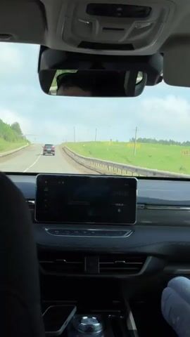 A video taken from inside a car driving on a road. The road is paved and appears to be a highway. There is a green grassy field on the right side of the road with trees in the distance. The sky is blue with some clouds. A black SUV is driving in front of the car. The interior of the car is visible, including the dashboard, steering wheel, and a rearview mirror. The video is shaky, indicating that the car is in motion. There are no people clearly visible in the video, other than a partial reflection of a person in the rearview mirror.