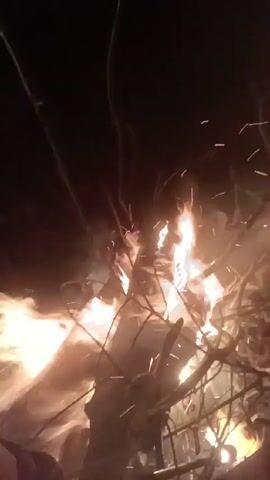 A video filmed at night shows a bonfire burning. Flames are visible, along with sparks. Thin tree branches are situated around the base of the fire. Music plays in the background.