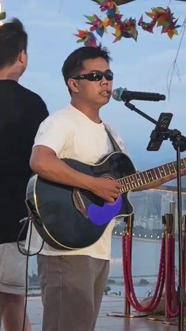 A man wearing sunglasses and a white t-shirt is playing an acoustic guitar and singing into a microphone. He is standing on a wooden stage, likely outdoors, with a cityscape and a body of water visible in the background. Colorful pinwheels are hanging above him. Another person in a black t-shirt and shorts stands behind him. The lighting suggests it is either early morning or late afternoon. Red ropes with gold stanchions are present, indicating a roped-off area, possibly a performance space or a viewing platform. A phone is mounted on a stand near the microphone, likely for lyrics or music. One can see some Chinese text on a sign in the far right of the image. The overall mood appears to be relaxed and enjoyable, suggesting a performance or a casual gathering.