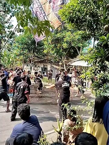A group of people in black clothing are participating in a cultural event on a street. Some are performing what appears to be a dance or ritual, with one person holding a banner or flag above them. Others are watching from the sides. The environment is outdoors with trees and houses visible in the background. It appears to be a sunny day.