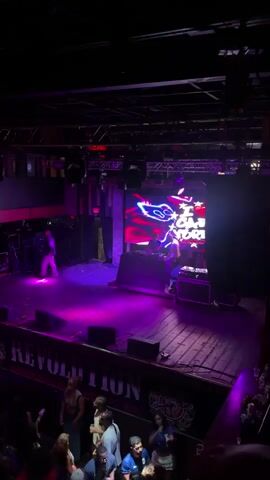 The video shows a crowded nightclub with a DJ on stage. The DJ booth is set up on a raised platform with a large LED screen behind it displaying a graphic. The screen has a red, white, and blue theme with stars and stripes, and some text that is difficult to read but appears to say "The Cash Back Tour". The stage is lit with purple and blue lights. In front of the stage, a crowd of people is dancing and socializing. The venue appears to be a medium-sized club with multiple levels, as indicated by the elevated perspective from which the video is filmed. The lighting is dim and atmospheric, with a mix of colored stage lights and general ambient light. The overall atmosphere is energetic and lively, typical of a nightclub setting.