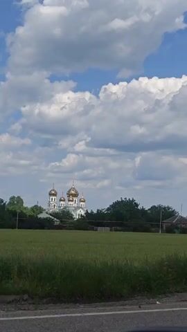 A white church with multiple gold domes stands behind a line of trees. A field of green crops is in the foreground, and a dark road is at the bottom of the frame. A triangular signpost is visible on the left side of the frame, and a small house is visible on the right. The sky is blue with large white clouds.
