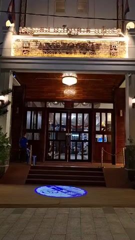 This video captures the exterior of a hotel or residence at night, likely in Sihanoukville, Cambodia. The entrance is well-lit, highlighting a prominent sign with a glowing illuminated border. The sign features text in what appears to be Khmer script and some English text that is partially visible as "THE HIDDEN PLACE". A circular blue light projection of a logo is cast on the wooden walkway leading to the entrance.

The architecture suggests a modern building with traditional influences, featuring dark wood paneling around the entrance and large glass doors. Two flags, one appearing to be the Cambodian flag, are visible flying near the top of the building. A single person, dressed in a blue shirt and dark pants, stands near the entrance to the left, interacting with their phone. Lush greenery, including palm trees and smaller potted plants, is visible around the entrance, illuminated by string lights, suggesting a tropical environment. The lighting, along with the building's facade, creates an inviting and somewhat luxurious atmosphere. The overall impression is one of a well-maintained and attractive establishment preparing to receive guests in the evening.