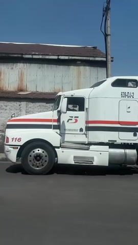 A white semi-truck and its attached trailer are seen moving slowly from left to right across the frame on a bright, sunny day with a clear blue sky. The truck's cabin is white with red and black horizontal stripes. On the side of the cabin, the numbers "639-DJ-2" and "116" are visible, along with a logo that appears to be "FS" or similar. The trailer is also white with red and black horizontal stripes, matching the truck. On the side of the trailer, the number "132" is displayed. In the background, there is a building with a corrugated metal roof. The ground is paved. The video captures the side profile of the vehicle as it passes by. The setting appears to be an outdoor, possibly industrial or commercial, area in Buenavista, Mexico.